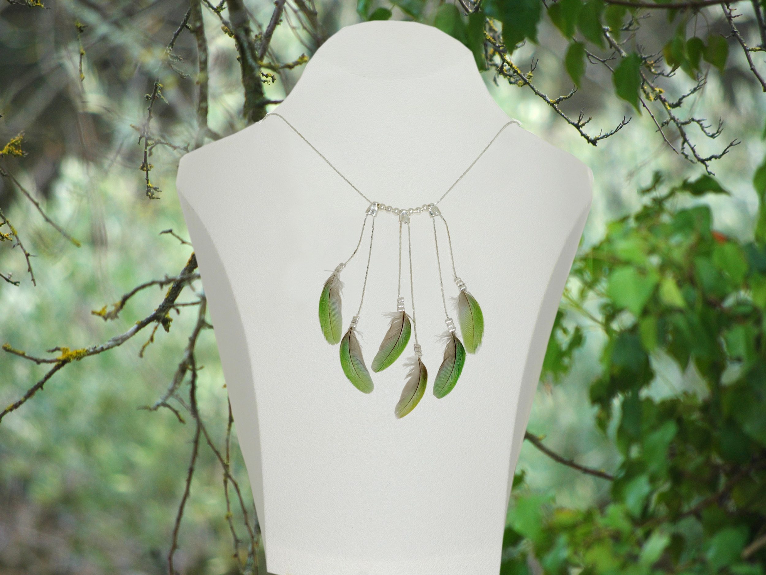 Un collier fait de chaînes argentées et de plumes vertes, présenté dans un décor végétal.
