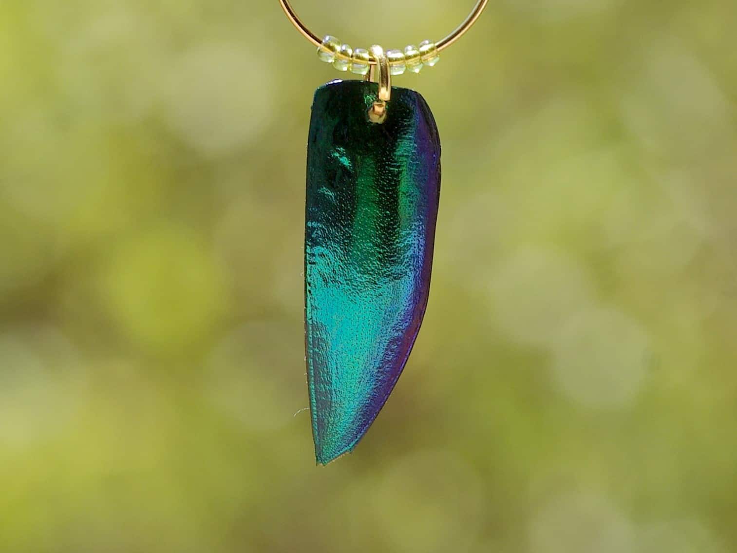 détail d'une boucles d'oreilles : élytre de scarabée bleu-vert accrochés à un anneau doré avec de petites perles vert clair,