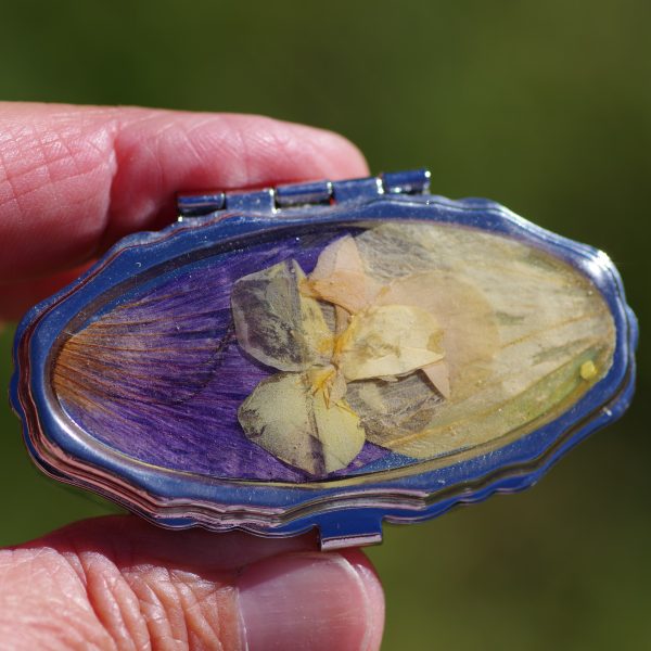petite boite à pilule ovale joliment décorée de fleurs , décor bicolore violet et jaune, avec une mini pensée jaune. Le boitier est argenté
