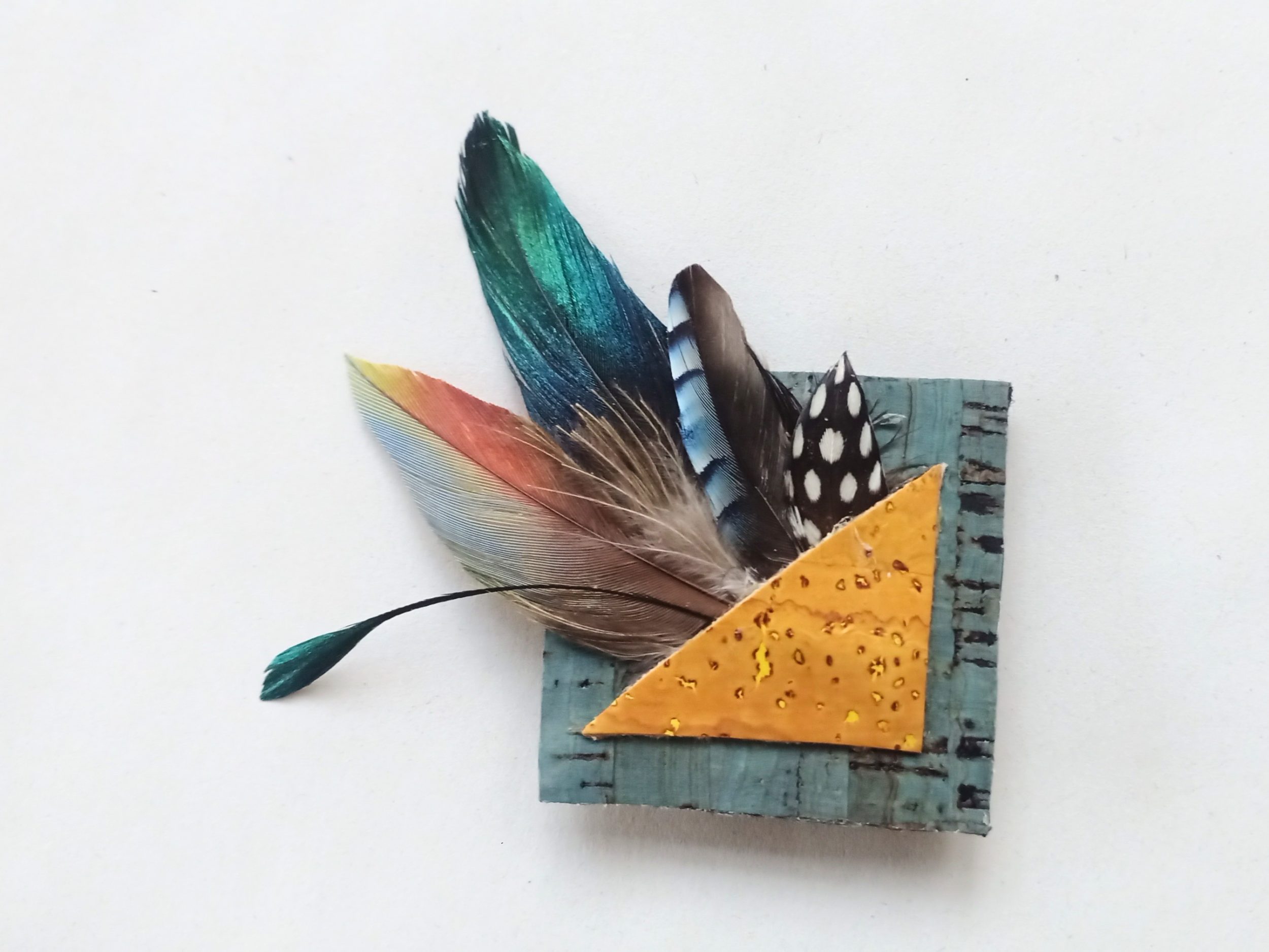 broche carrée avec une composition de plumes et liège : triangle jaune dans carré bleu et explosion de plumes multicolores