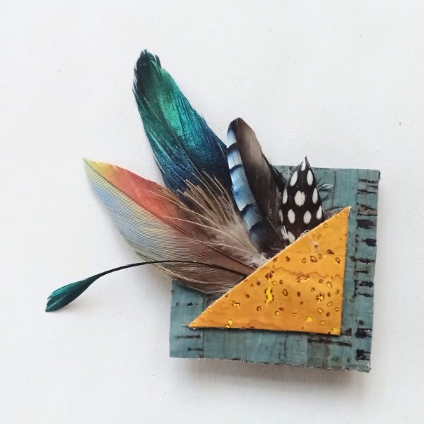 broche carrée avec une composition de plumes et liège : triangle jaune dans carré bleu et explosion de plumes multicolores