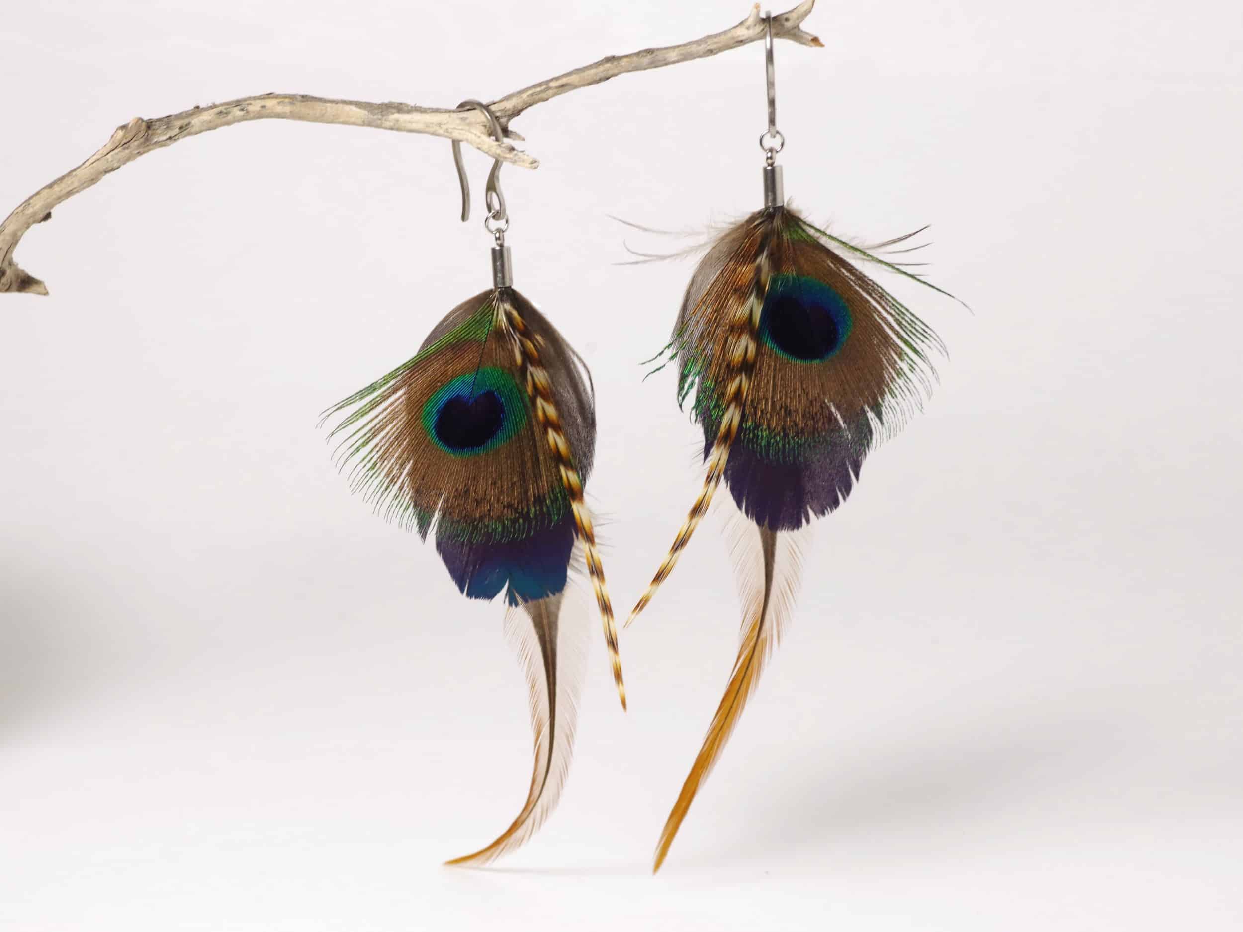 Une paire de boucles d'oreilles avec des plumes longues et effilées dans des tons kaki, bleu et brun (dont un œil de paon).
