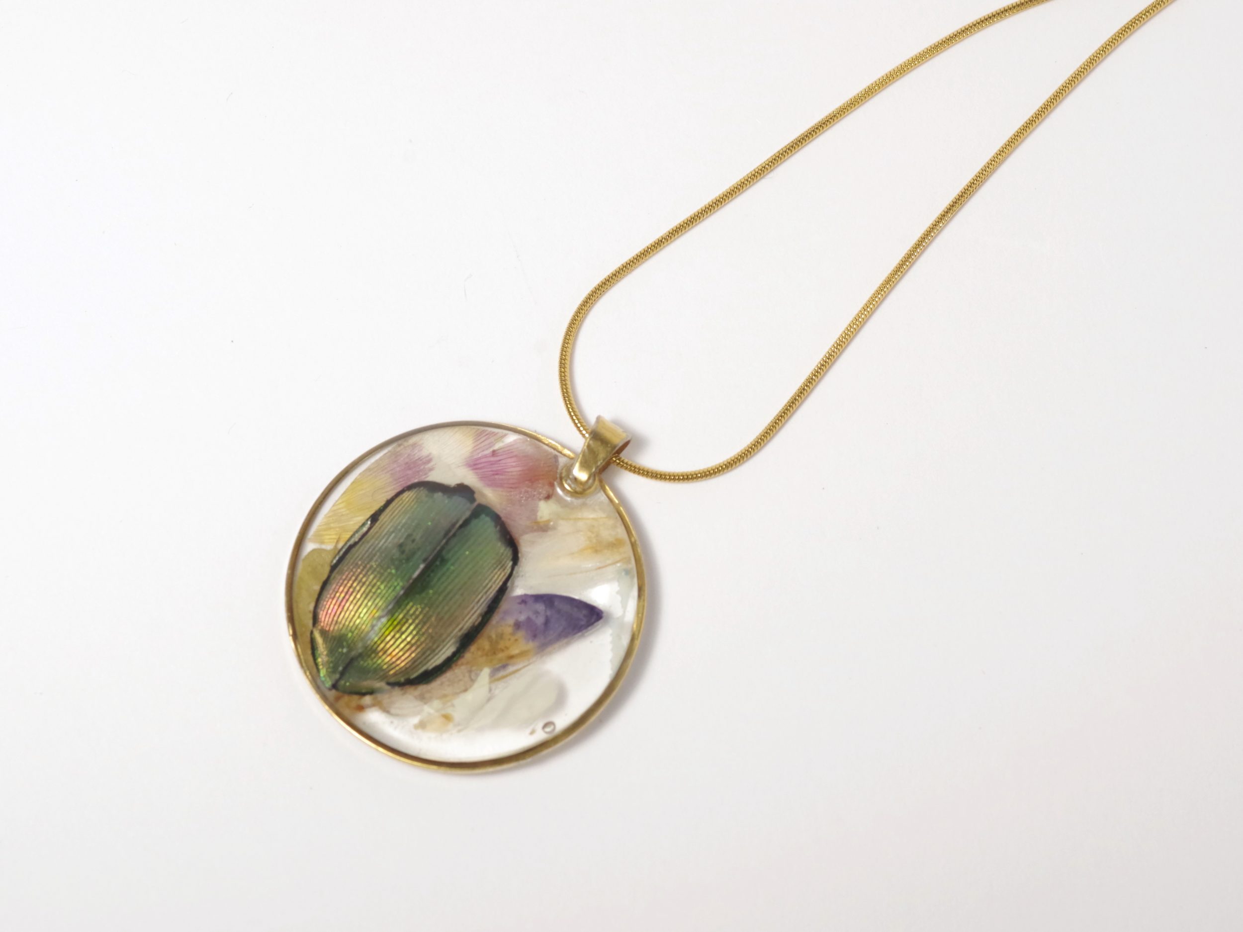 Un pendentif avec un petit scarabée aux couleurs irisées, inclus dans de la résine avec des fleurs des champs.