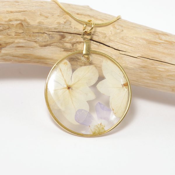 Un pendentif avec deux fleurs blanches et une petite fleur violette coulées dans une résine transparente dans un cercle en inox doré.