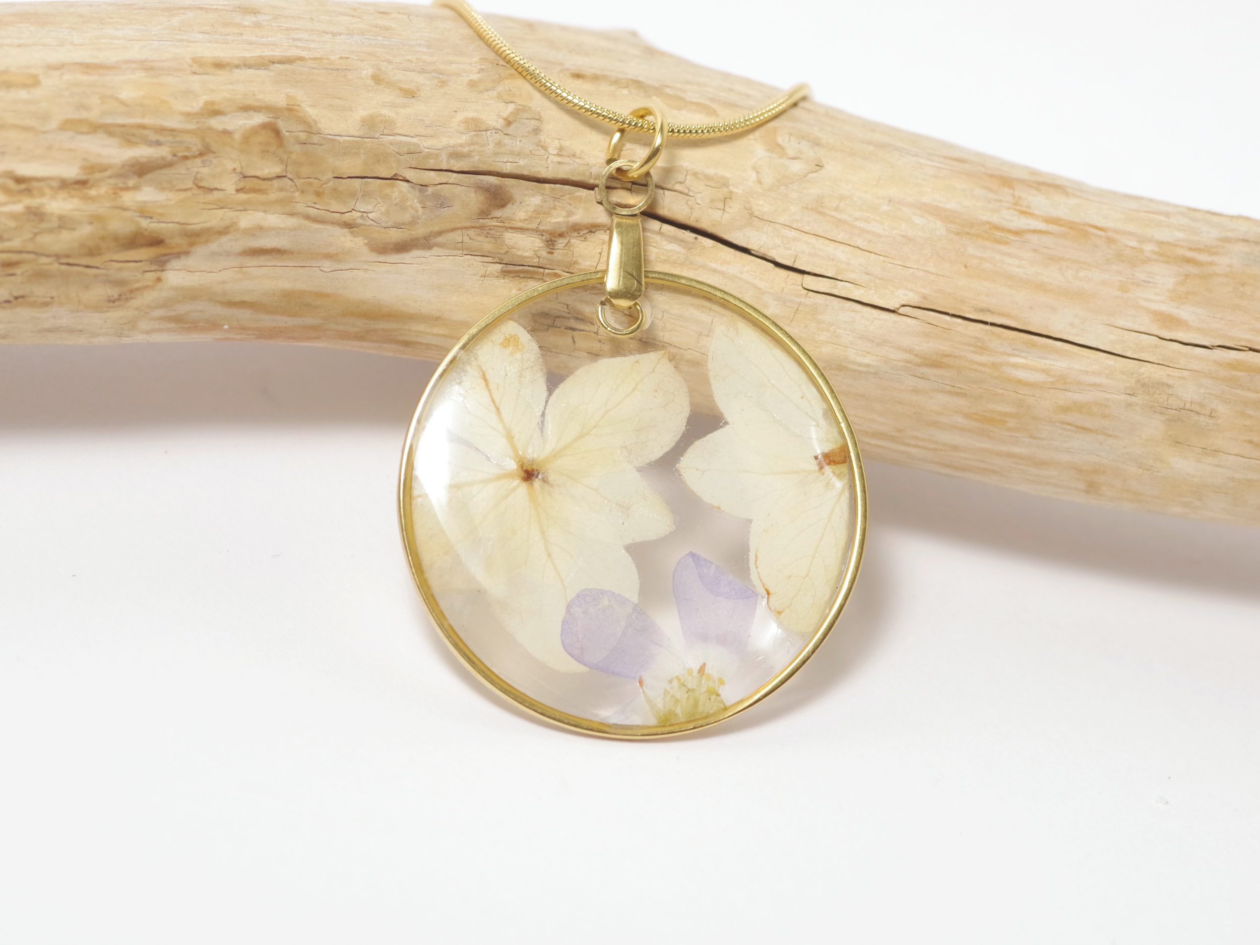 Un pendentif avec deux fleurs blanches et une petite fleur violette coulées dans une résine transparente dans un cercle en inox doré.