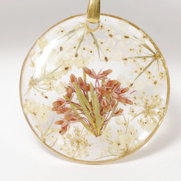 Pendentif fleuri : minis fleurs banc et rose coulées dans de la résine dans un fin cadre doré rond.
